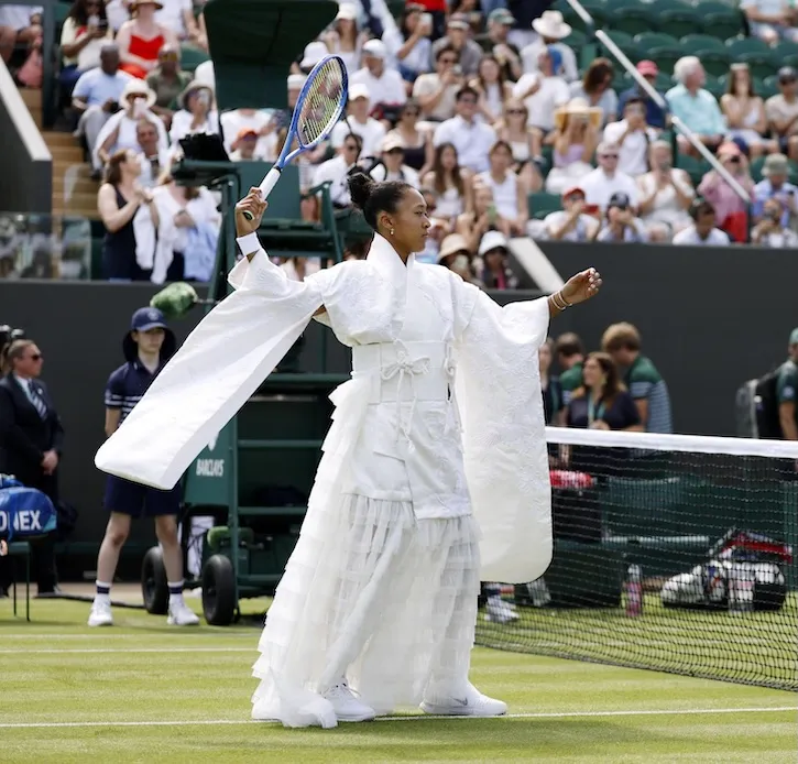 Naomi Osaka op Wimbledon met een witte geborduurde kimono, wijde mouwen en tennisracket in de hand